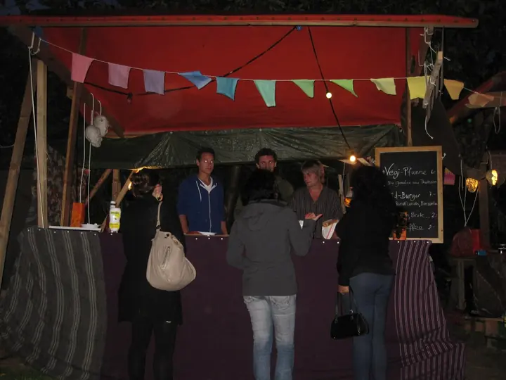 Festivalgäste stehen nachts an einem rot geschmückten Holzstand.