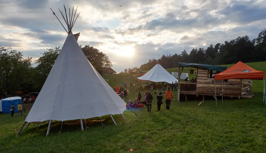 Ein Tipi und Holzbauten auf der Festivalwiese im Abendlicht.