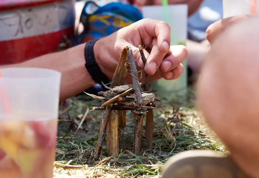Hände bauen auf einer Wiese eine kleine Konstruktion aus Zweigen.
