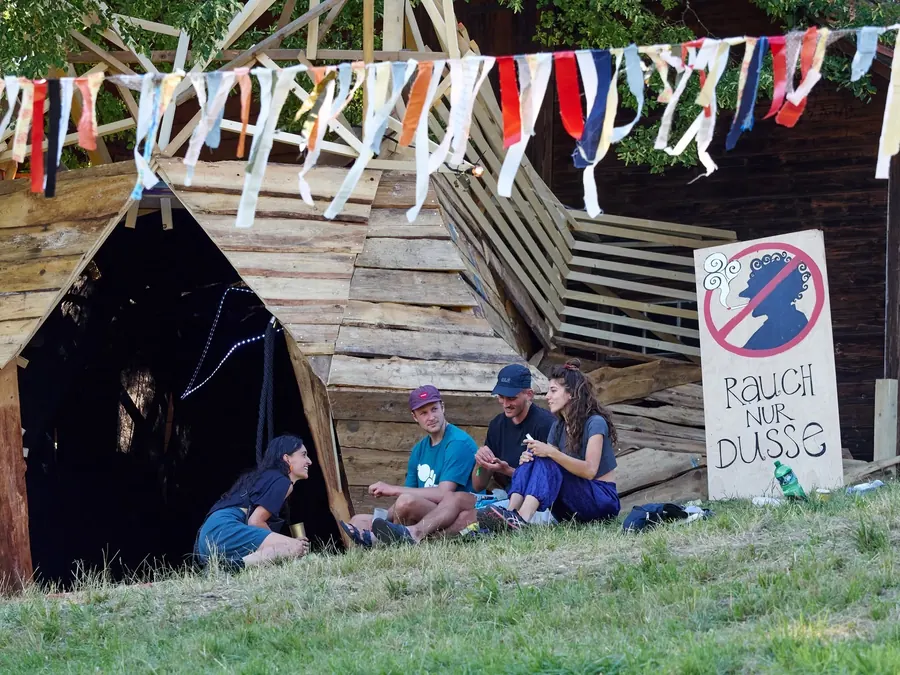 Festivalgäste sitzen im Schatten eines selbstgebauten Holzunterstands.